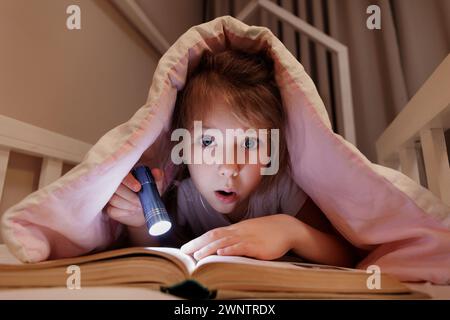 surprised girl lying in bed and reading book with flashlight under blanket in dark room Stock Photo