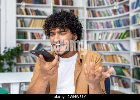 A young man with curly hair appears exasperated while having a heated conversation on his smartphone, standing against a backdrop of bookshelves in a library setting. Stock Photo