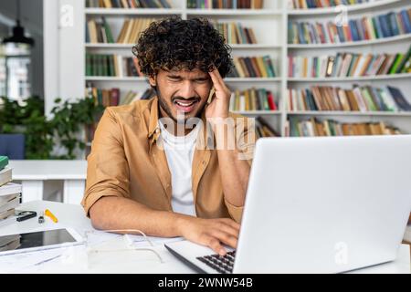 A young man with curly hair is visibly stressed and experiencing a headache as he works on his laptop, possibly dealing with work pressure or studying challenges in a home environment Stock Photo