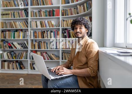 An engaged young adult sits comfortably in a well-lit space, surrounded by books, focused on work being done on a laptop. Setting suggests a modern library or a study room with a peaceful atmosphere. Stock Photo