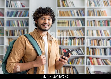 A cheerful young man with curly hair and a backpack is standing in a library. He is casually dressed, smiling,using a smartphone, possibly searching for information or communicating with friends. Stock Photo