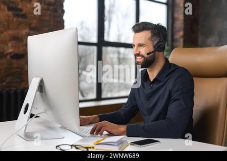 A charismatic businessman wearing headphones and a smart casual shirt is sitting at a desk in front of a personal computer, involved virtual meeting Stock Photo