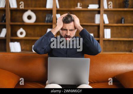 An unhappy young male freelancer looks at a laptop screen shocked by gadget breakdown or operational problems. A frustrated man confused and surprised by an unexpected error on a computer device Stock Photo