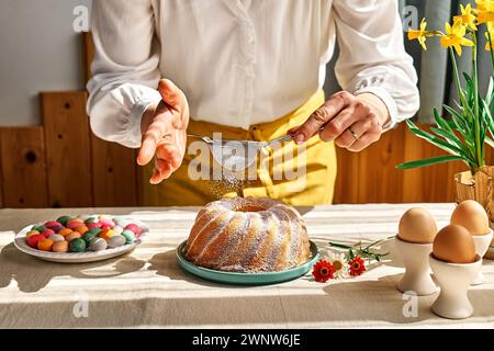 Female hands sprinkles Easter cake with icing sugar through sieve. Anonymous woman making traditional easter cake or sweet bread with topping. Easter Stock Photo