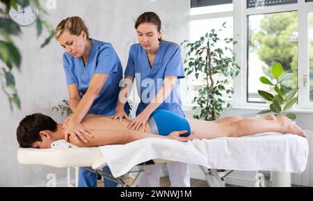 Two female masseuses perform stroking and rubbing of muscles of back and shoulders to guy client Stock Photo