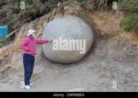 A woman points to a Moeraki boulder on Koekohe beach on the South Island of New Zealand. The large boulders, on the Otago coast, are unusually round. Stock Photo