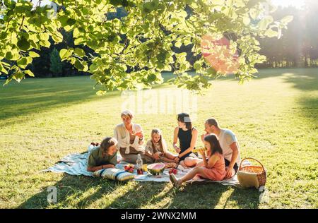 Big family under Linden tree on the picnic blanket on the in city park green grass. They are eating boiled corn, apples, peaches, pastries and waterme Stock Photo