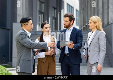 A group of business professionals engaged in a discussion outside office buildings. They are dressed smartly in suits, and one is holding a digital tablet. Stock Photo