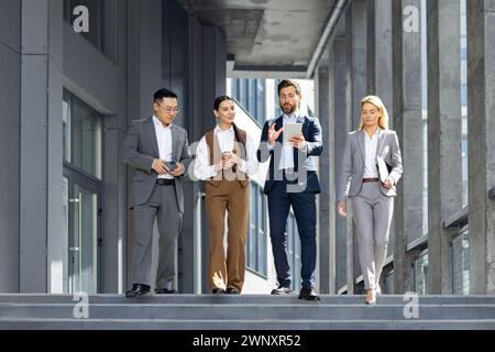 A group of four business professionals confidently strolling down steps outside a modern office building, engrossed in discussion with digital tablets and smartphones in hand. Stock Photo