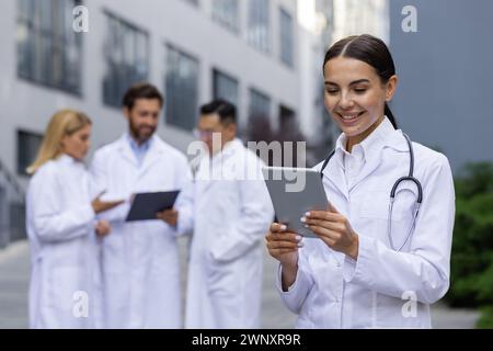A professional female healthcare worker with a stethoscope around her neck is using a tablet outdoors. In the blurred background, her colleagues are having a discussion. Stock Photo