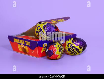 Opened box of opened Cadbury Creme Eggs against a light purple background with two creme eggs in front Stock Photo