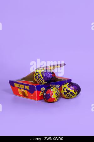 Opened box of opened Cadbury Creme Eggs against a light purple background with two creme eggs in front Stock Photo
