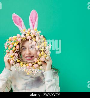 Happy Easter. Girl with joyful smile, in shiny pink rabbit ears and holding an Easter wreath of flowers, against blue background. He smiles sincerely Stock Photo