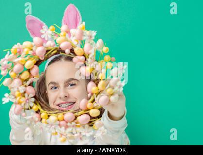 Happy Easter. Girl with joyful smile, in shiny pink rabbit ears and holding an Easter wreath of flowers, against blue background. He smiles sincerely Stock Photo