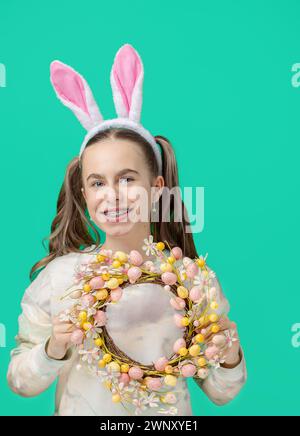 Happy Easter. Girl with joyful smile, in shiny pink rabbit ears and holding an Easter wreath of flowers, against blue background. He smiles sincerely Stock Photo