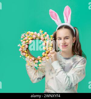 Happy Easter. Girl with joyful smile, in shiny pink rabbit ears and holding an Easter wreath of flowers, against blue background. He smiles sincerely Stock Photo