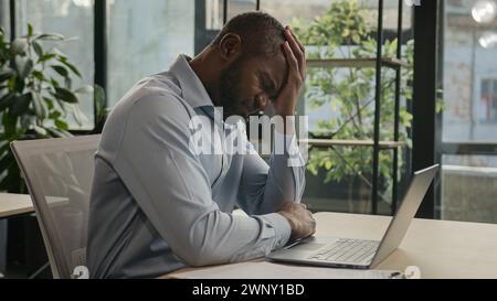 Depressed African american ethnic mature businessman stressful sick man sit in office with laptop read papers documents business accounting worried of Stock Photo