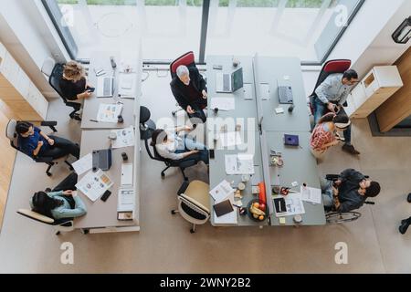 Overhead view of a collaborative workspace with a group of people engaged in discussion, working on computers and brainstorming around a shared table. Stock Photo