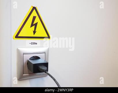 High-voltage sign on the white wall. A plug with a cable is stuck in an electrical outlet. Charging your phone or smartphone. The danger of electric Stock Photo