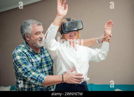 Immersed in the wonders of modern technology, a senior couple enjoys a playful moment in their living room. The woman wears virtual reality glasses, t Stock Photo