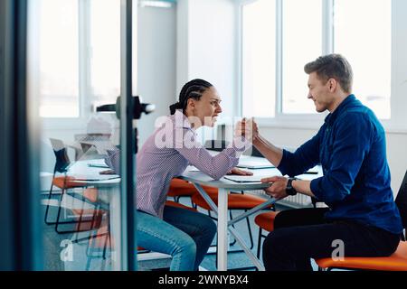 In a modern startup office, a businessman and a businesswoman business colleagues engage in a symbolic arm-wrestling match, reflecting teamwork Stock Photo