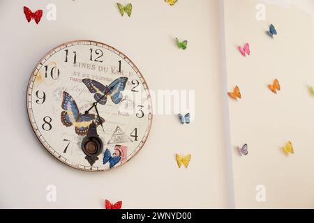 Wall clock with butterfly designs hangs on a beige wall with colorful butterfly figures Stock Photo