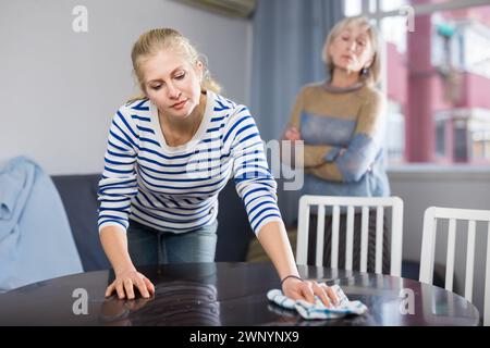 Elderly mother is unhappy with the way her adult daughter dusts the table in room Stock Photo