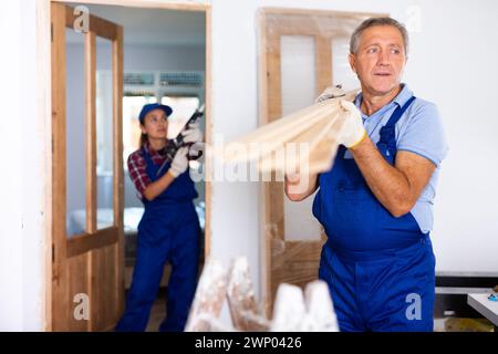 Confident middle-aged repairman in uniform carrying wooden timber at a construction site in an apartment Stock Photo