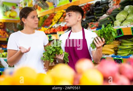 Doubted young female customer asking advice from male market assistant while buying greens in supermarket Stock Photo