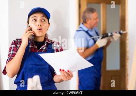 Worried Latin female repairer in uniform talking on a mobile phone while holding a work plan while renovating a house Stock Photo