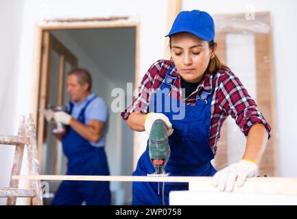 Focused Latin female in uniform using a jigsaw machine on wooden plank at home Stock Photo
