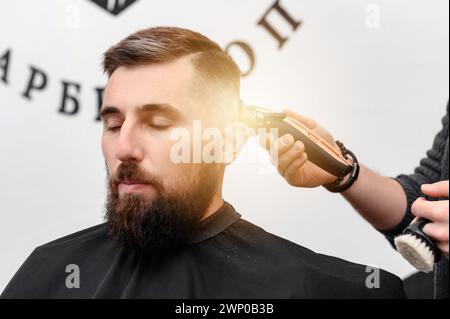 A barber cuts a man with a beard in a barber shop. Short haircut of the client with a clipper. Stock Photo