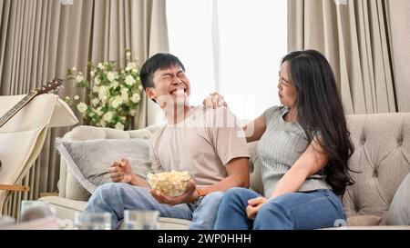 A cheerful and joyful young Asian couple is laughing at jokes and enjoying popcorn while watching television together at home. couple and domestic lif Stock Photo