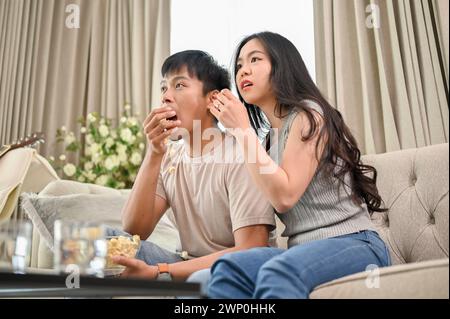 An excited, shocked young Asian couple is watching an exciting TV show or movie on a sofa together at home. couple and domestic life concepts Stock Photo