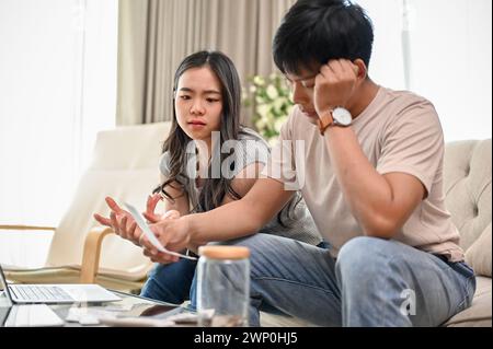 An unhappy and stressed young Asian couple is having a dispute over their household expenses and finances, arguing about high domestic bills on a sofa Stock Photo