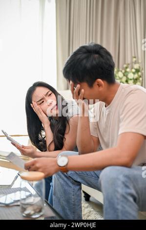 An unhappy and stressed young Asian couple is having a dispute over their household expenses and finances, arguing about high domestic bills on a sofa Stock Photo
