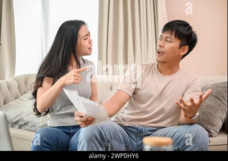 A resentful, angry Asian wife and husband are arguing about family household invoices and high domestic bills, quarreling about the issue of wasting m Stock Photo