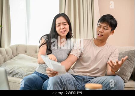 A resentful, angry Asian wife and husband are arguing about family household invoices and high domestic bills, quarreling about the issue of wasting m Stock Photo