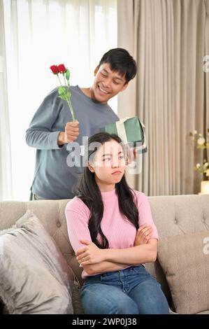 A romantic Asian husband is trying to ask for forgiveness from his angry wife by surprising her with a present and flowers in the living room. family Stock Photo