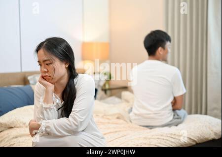 An unhappy, sad young Asian couple in pajamas is sitting separately on the bed after their argument, not talking to each other, having problems in the Stock Photo