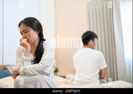 An unhappy, sad young Asian couple in pajamas is sitting separately on the bed after their argument, not talking to each other, having problems in the Stock Photo