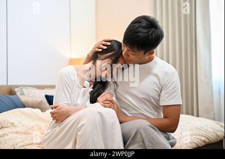 A caring Asian man is hugging his sad girlfriend, apologizes, asking for forgiveness, and making peace after quarrel in the bedroom. relationship prob Stock Photo