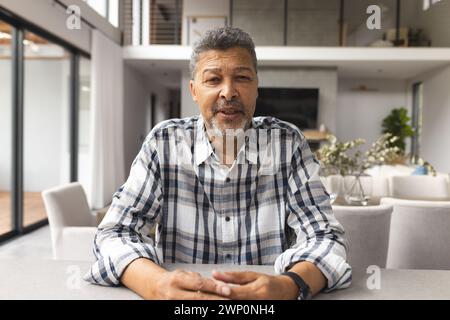 Biracial senior man with salt-and-pepper hair sits at a table on a video call, smiling gently Stock Photo