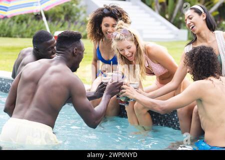 Diverse group of friends enjoy a pool party, toasting drinks with joyful expressions Stock Photo