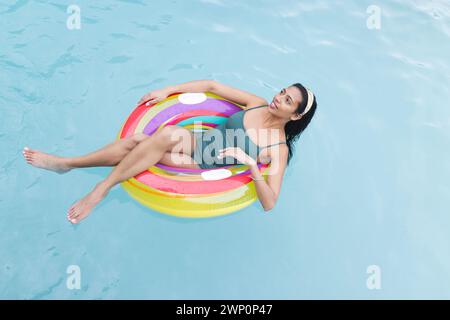 Young biracial woman relaxes on a colorful pool float, her legs gently dipped in the water Stock Photo