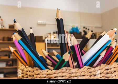 Multicolored wooden pencils with sharpened tips placed together in wicker basket against blurred shelves and interior of room in daylight Stock Photo