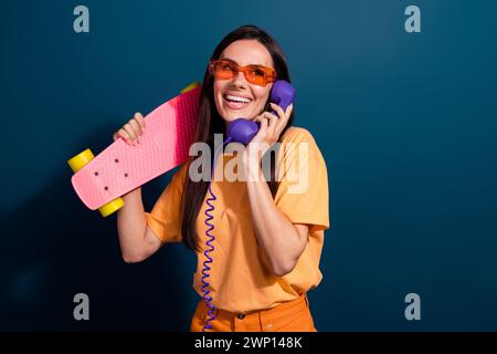 Portrait of hipster woman wearing orange t shirt call old phone invite friends ride skateboard isolated on dark blue color background Stock Photo