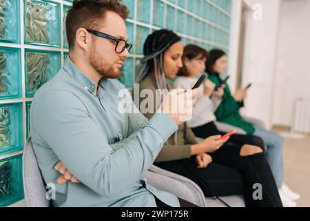 Side view of focused young diverse ethnicity candidates using mobile phones while waiting job interview in modern office lobby. Bored male and female Stock Photo