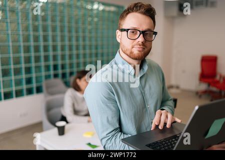 Portrait of confident startup businessman in casual clothes and glasses holding laptop in hand standing in office, looking at camera. Diverse business Stock Photo