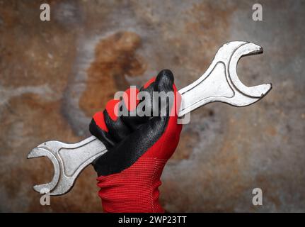 Close-up of a hand in red protective glove holding wrench against rusty metal background presenting a symbol of manual labor and repair Stock Photo